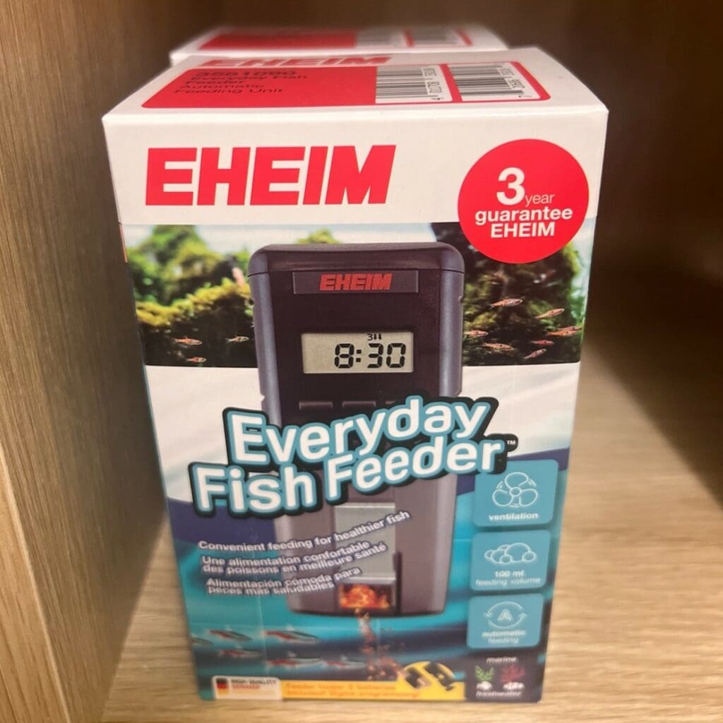 A boxed eheim everyday automatic fish feeder is displayed on a wooden shelf. The box highlights features such as automatic feeding, ventilation, and a 3-year guarantee. The digital feeder is shown on the packaging with a sample time of 8:30.