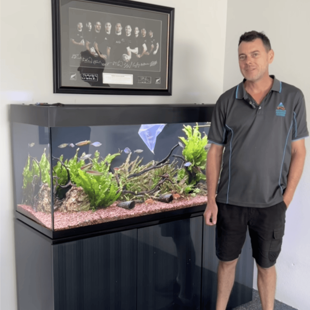 A man stands next to a large aquarium filled with plants and colorful fish, showcasing his dedication to aquarium maintenance. The tank sits on a black cabinet, with a signed photo hanging above. He wears a dark polo shirt and shorts.